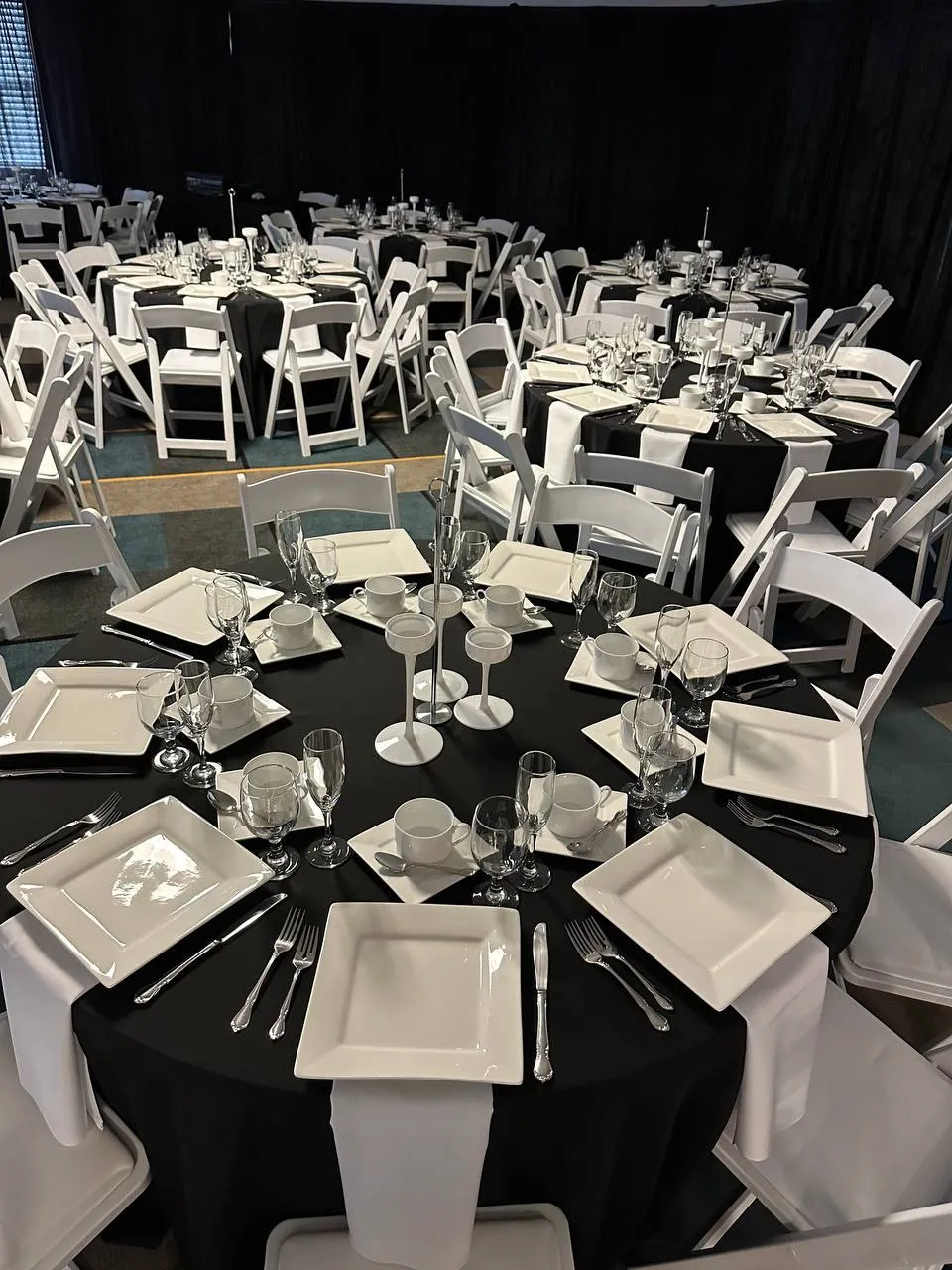 Black-draped round table set with square white plates, cups and saucers, glasses and silverware, with white chairs around it