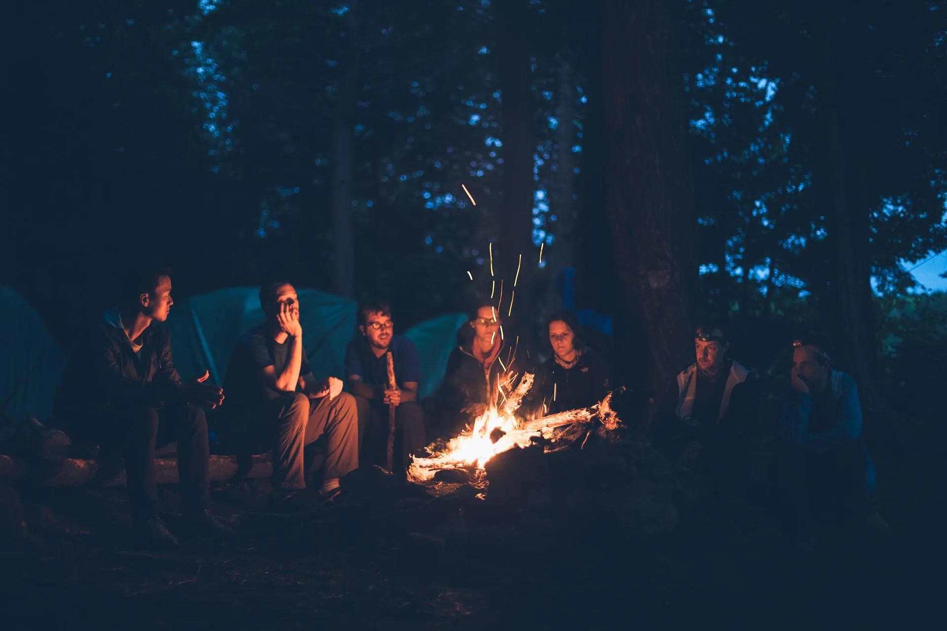 Group of people sitting around a campfire at night in a forest, with tents behind them and sparks rising