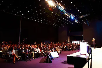 Large audience seated in a dark conference hall facing a speaker on a lit stage, with pinpoint lights across the ceiling