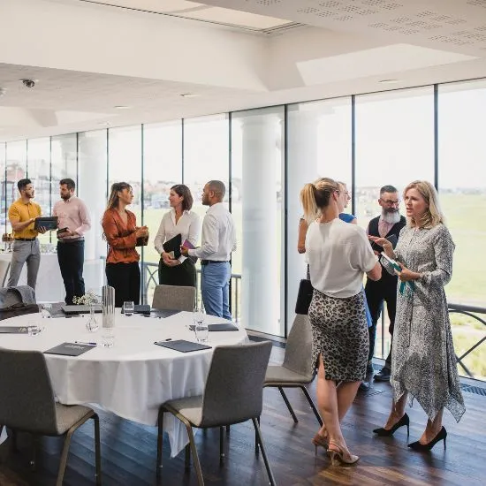 People in business attire mingling in a bright room with floor-to-ceiling windows, a white-linen round table and gray chairs