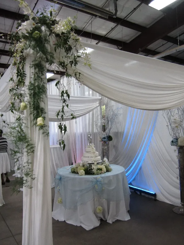 White draped fabric canopy with a floral garland over a round table holding a tiered cake, with blue uplighting behind