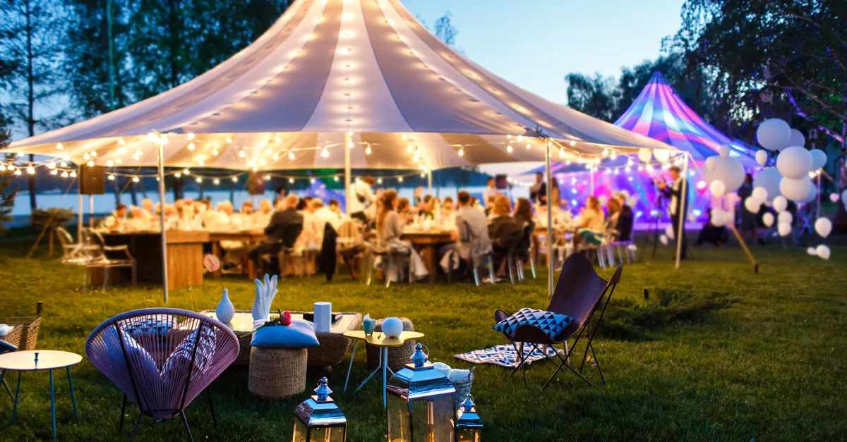 Dusk party under a string-lit white tent with guests at long tables, lanterns and lounge chairs on the lawn, a colorfully lit tent behind