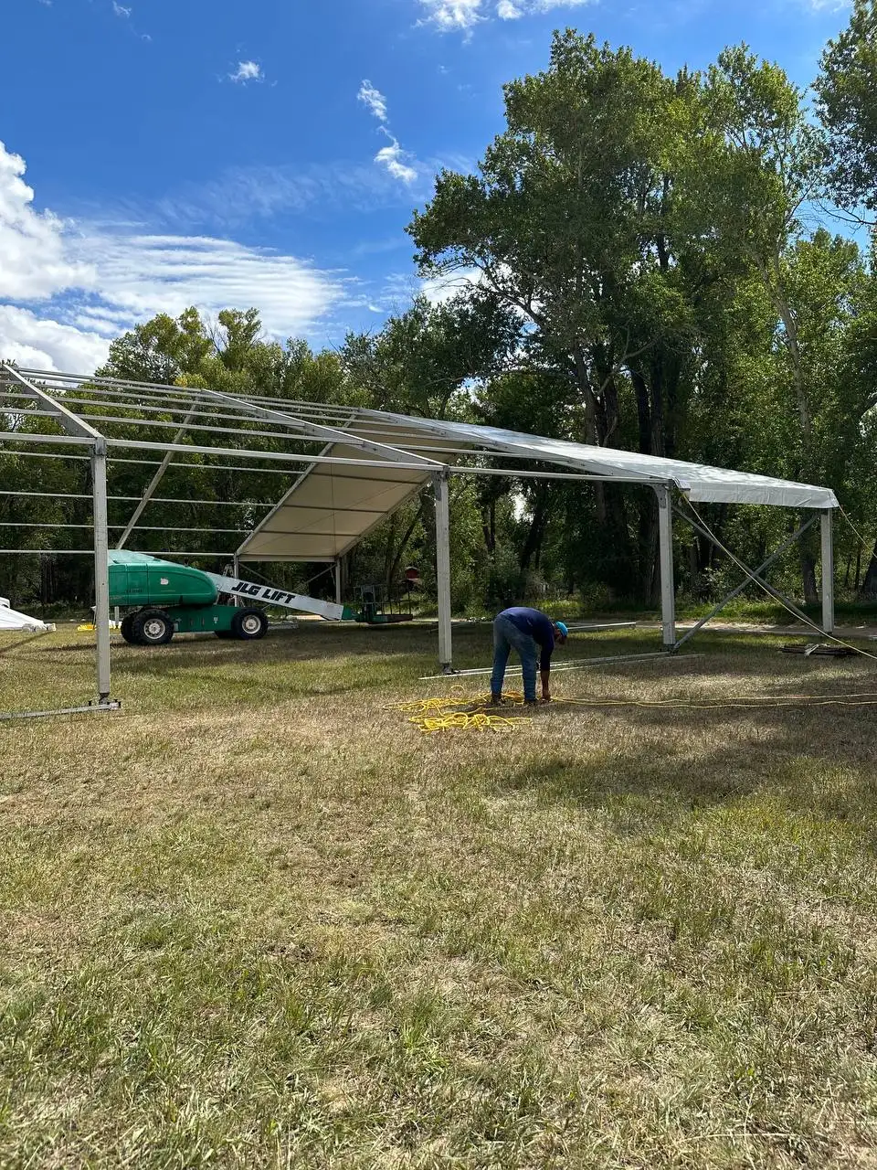 Frame tent going up on a grass lot: a worker at its edge and a lift beside the partly covered frame, tall trees behind