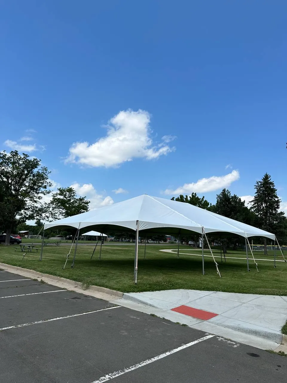White frame tent on a park lawn beside a parking lot, under a blue sky