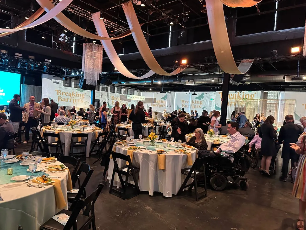Gala in a large hall with cream fabric swags across the ceiling, round tables with gold napkins and guests mingling