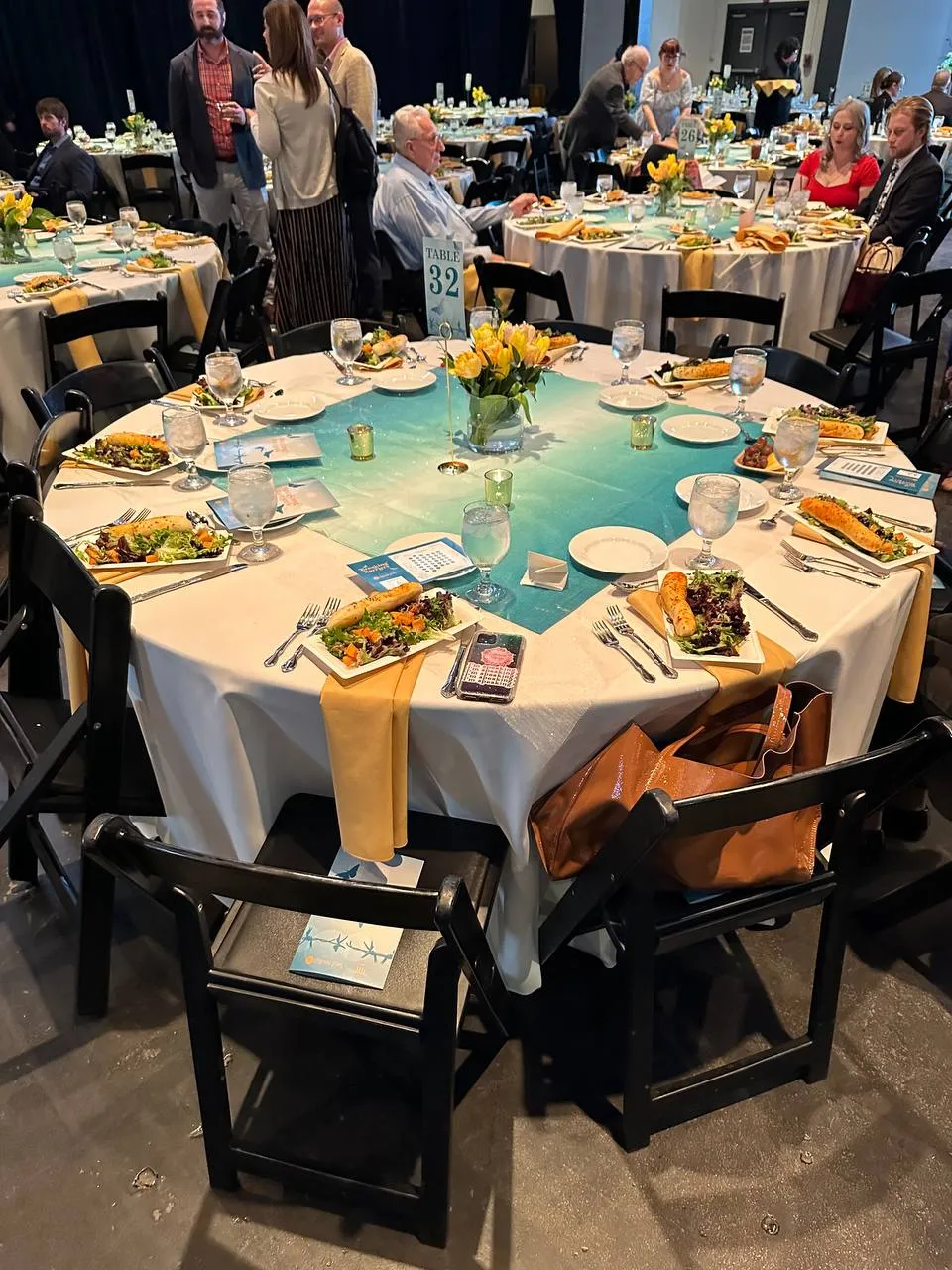 Round gala table with white linen, gold napkins, a teal runner, yellow tulips and plated salads, with guests nearby