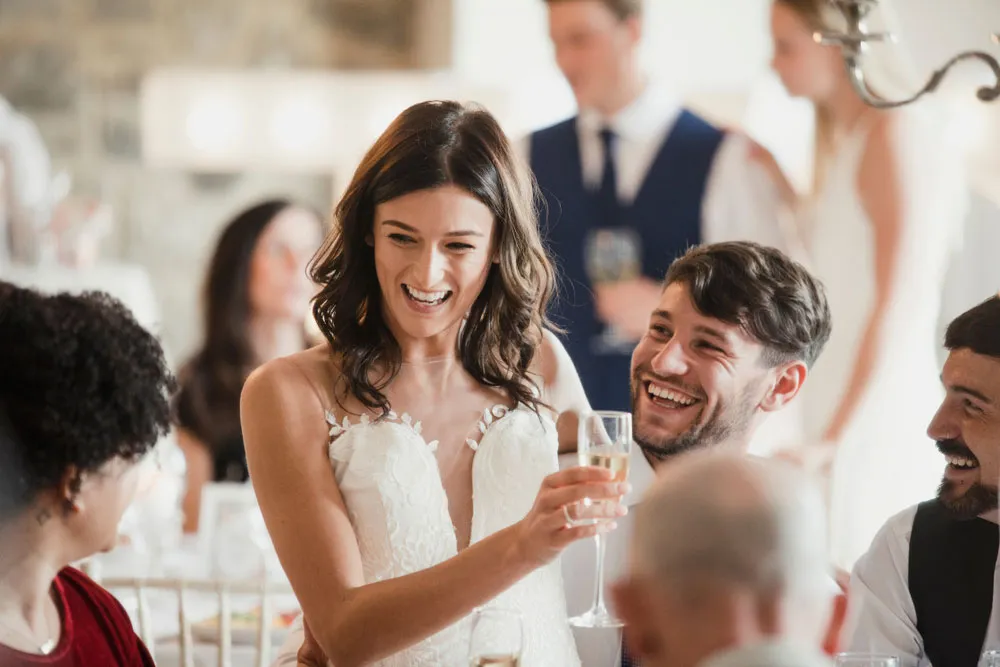 Smiling woman in a white lace gown raising a champagne glass among laughing guests seated at a table