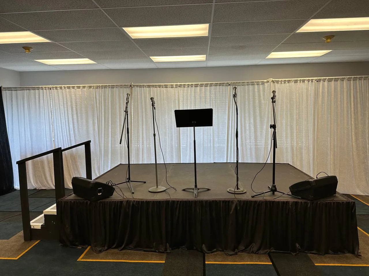 Indoor stage with black skirting, stairs and a handrail, microphone stands, a music stand and floor monitors before white drapes