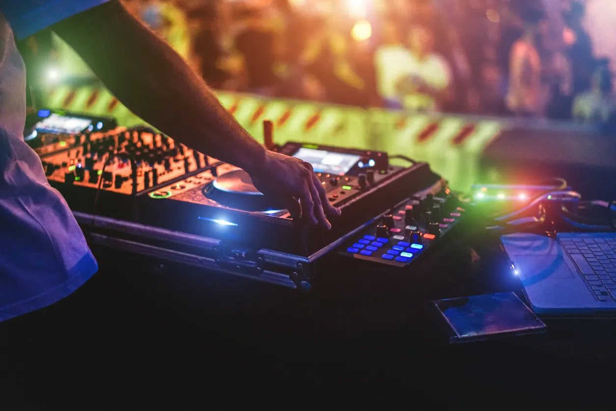 Hand on a DJ controller with a jog wheel, mixer knobs and blue-lit buttons, beside a laptop, with a blurred crowd behind