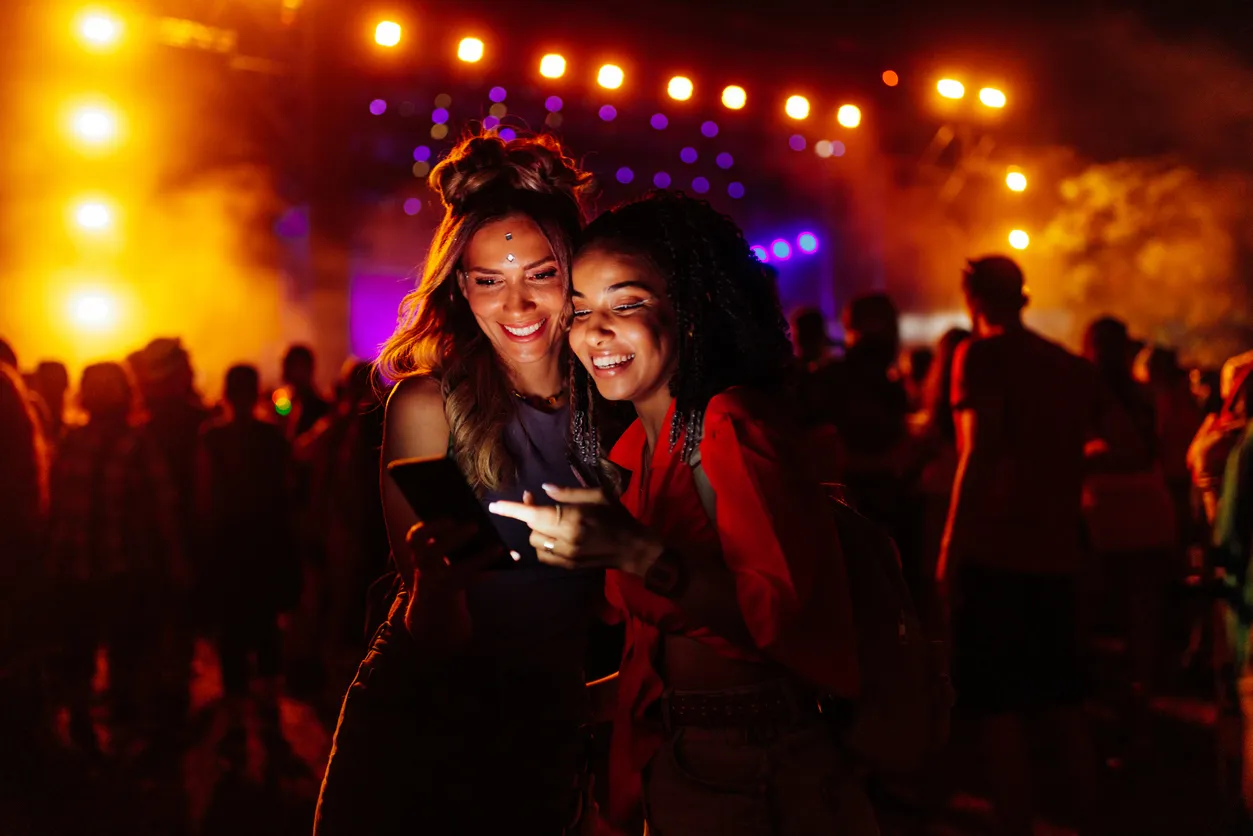 Two smiling women looking at a phone in a night crowd, with stage lights and haze glowing behind them