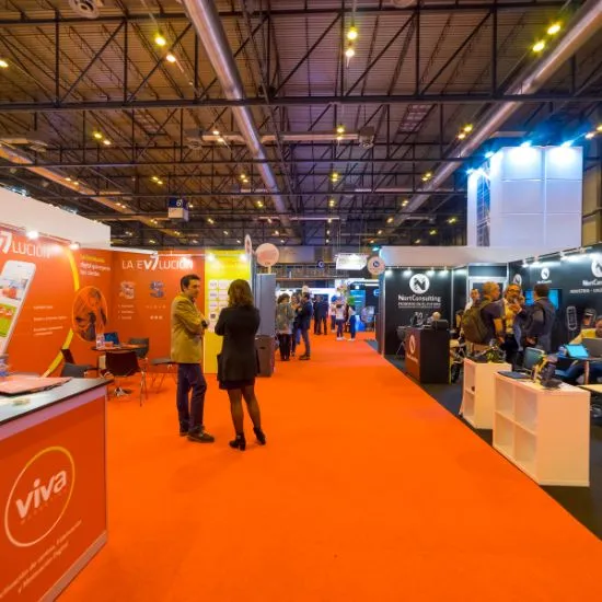 Exhibition hall aisle with bright orange carpet, booths on both sides and visitors browsing under a high truss ceiling