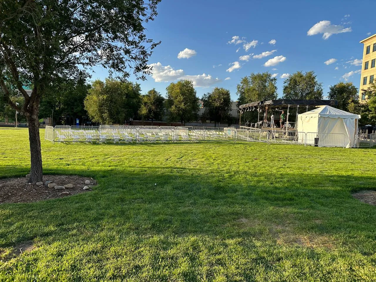 Rows of white folding chairs inside bike-rack barricades on a lawn, with a covered stage and a small white tent beyond