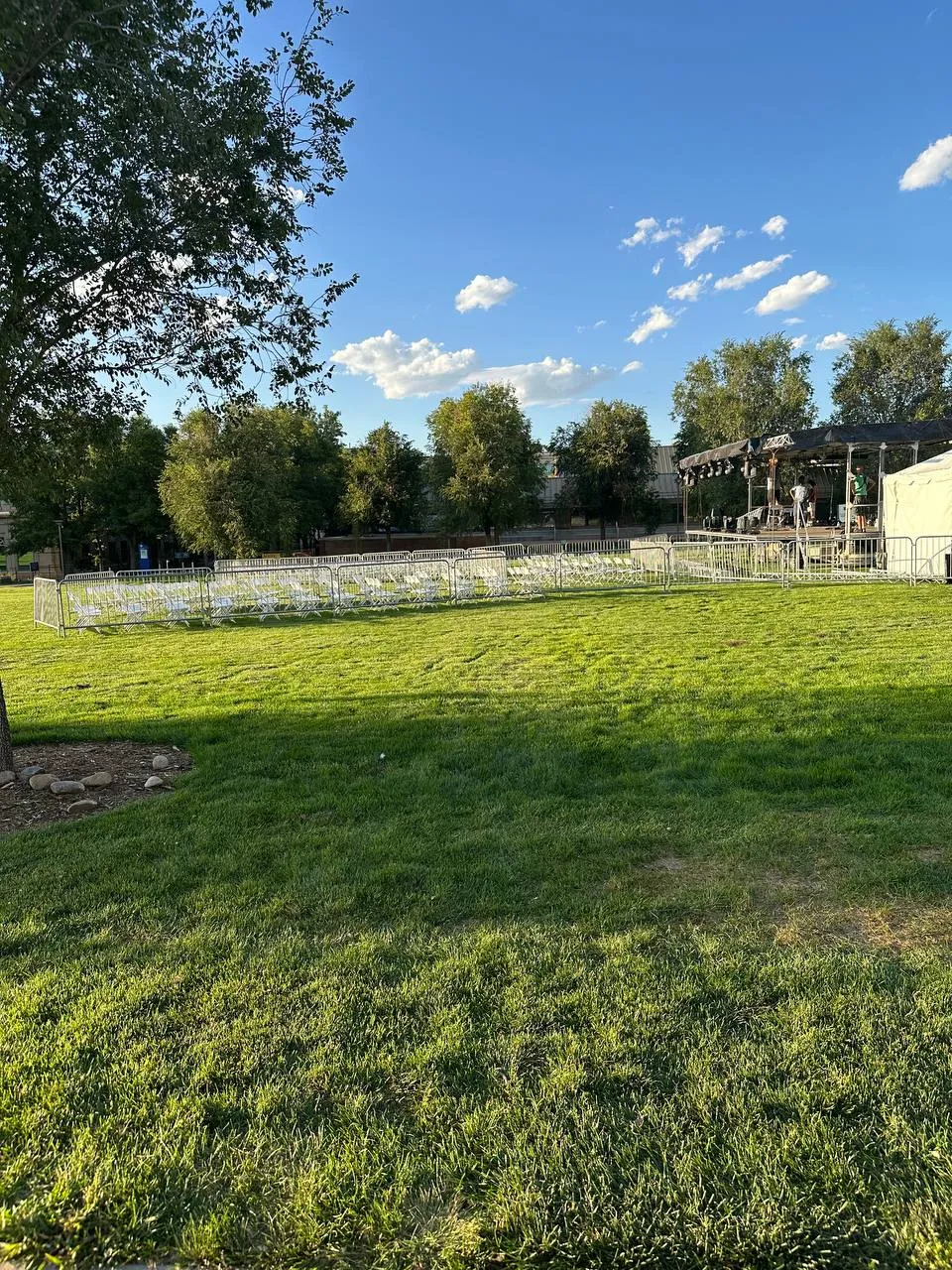 Rows of white chairs behind bike-rack barricades and a stage beyond a sunny lawn, with trees and a blue sky