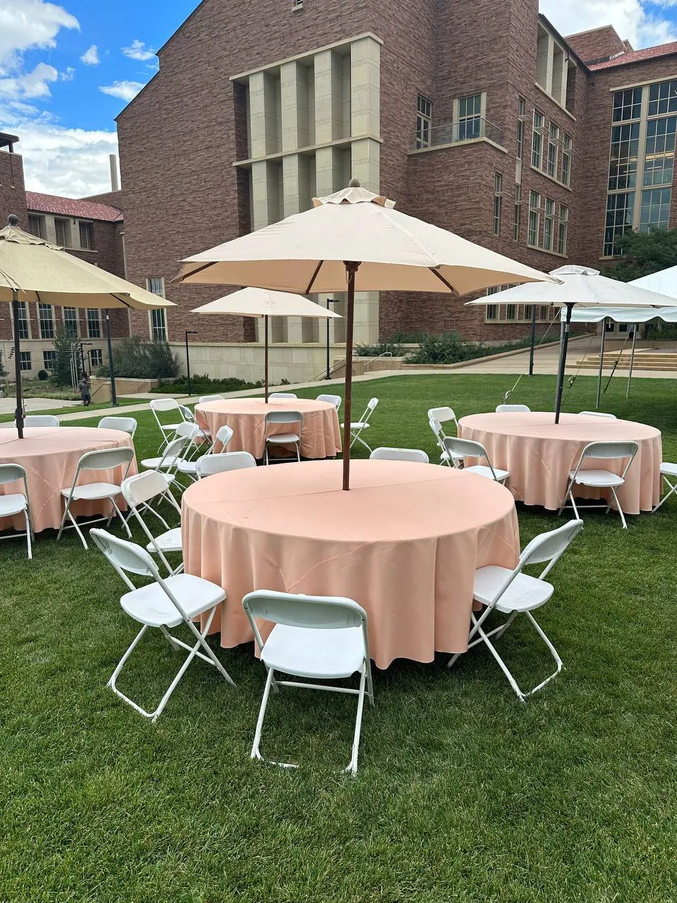 Round tables with blush tablecloths and white folding chairs under market umbrellas on a lawn by a brick building