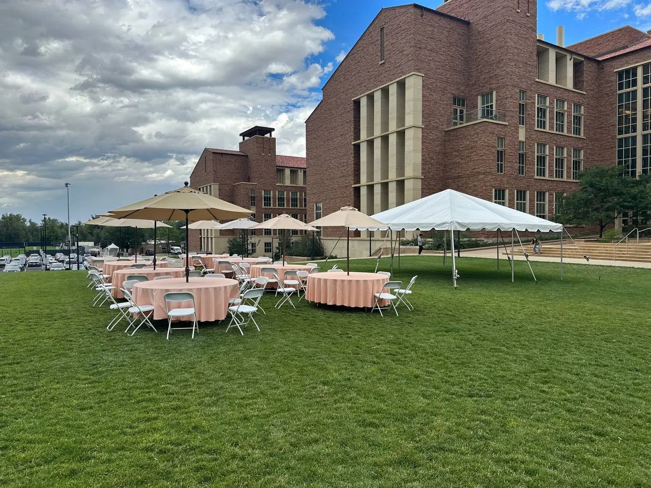 Round tables with blush linens, white folding chairs, market umbrellas and a white frame tent on a lawn beside brick buildings