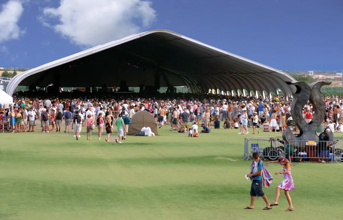 Huge curved-roof tent with stage lighting inside, a large crowd on the grass in front and a blue sky above