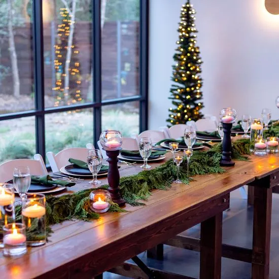 Long wooden farm table set with an evergreen garland, floating candles, glassware and green napkins beside a lit Christmas tree
