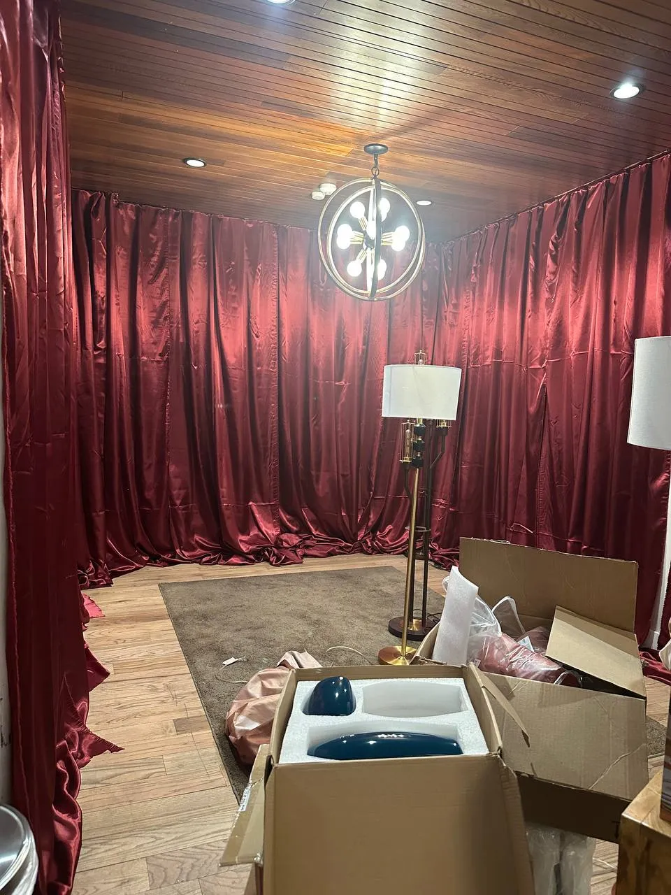 Small room lined with red satin drapes, with a ring chandelier, a floor lamp and boxes during setup