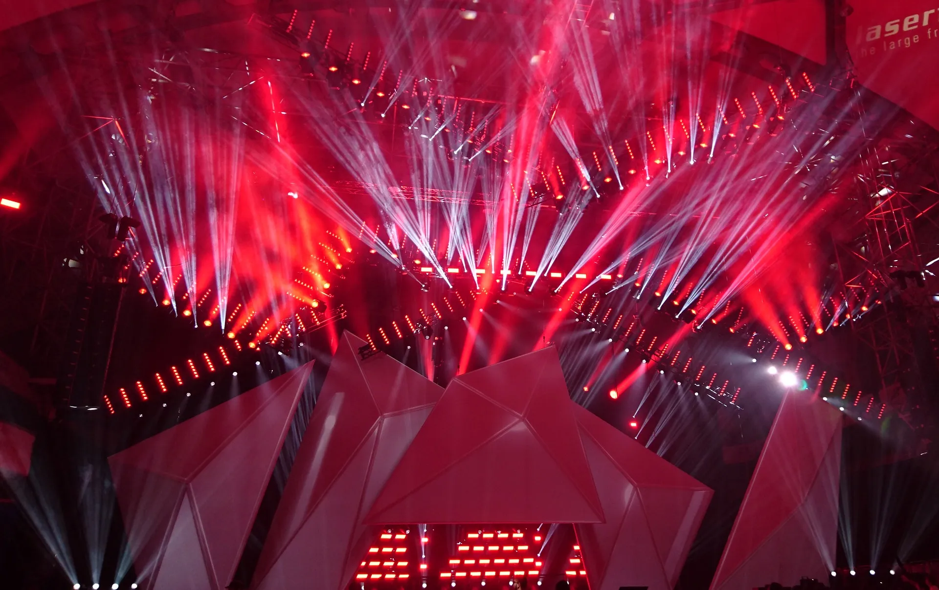 Stage lit in red with fanning white light beams, curved rows of LED lights and angular white set pieces in front
