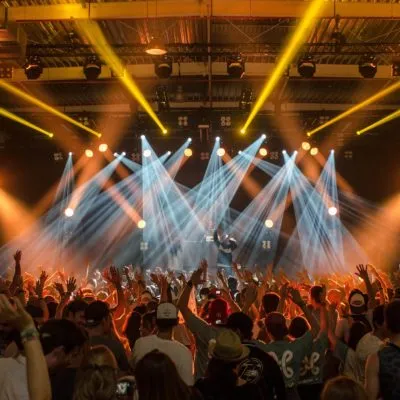 Concert crowd with raised hands facing a stage lit by white and yellow light beams under overhead truss and spotlights