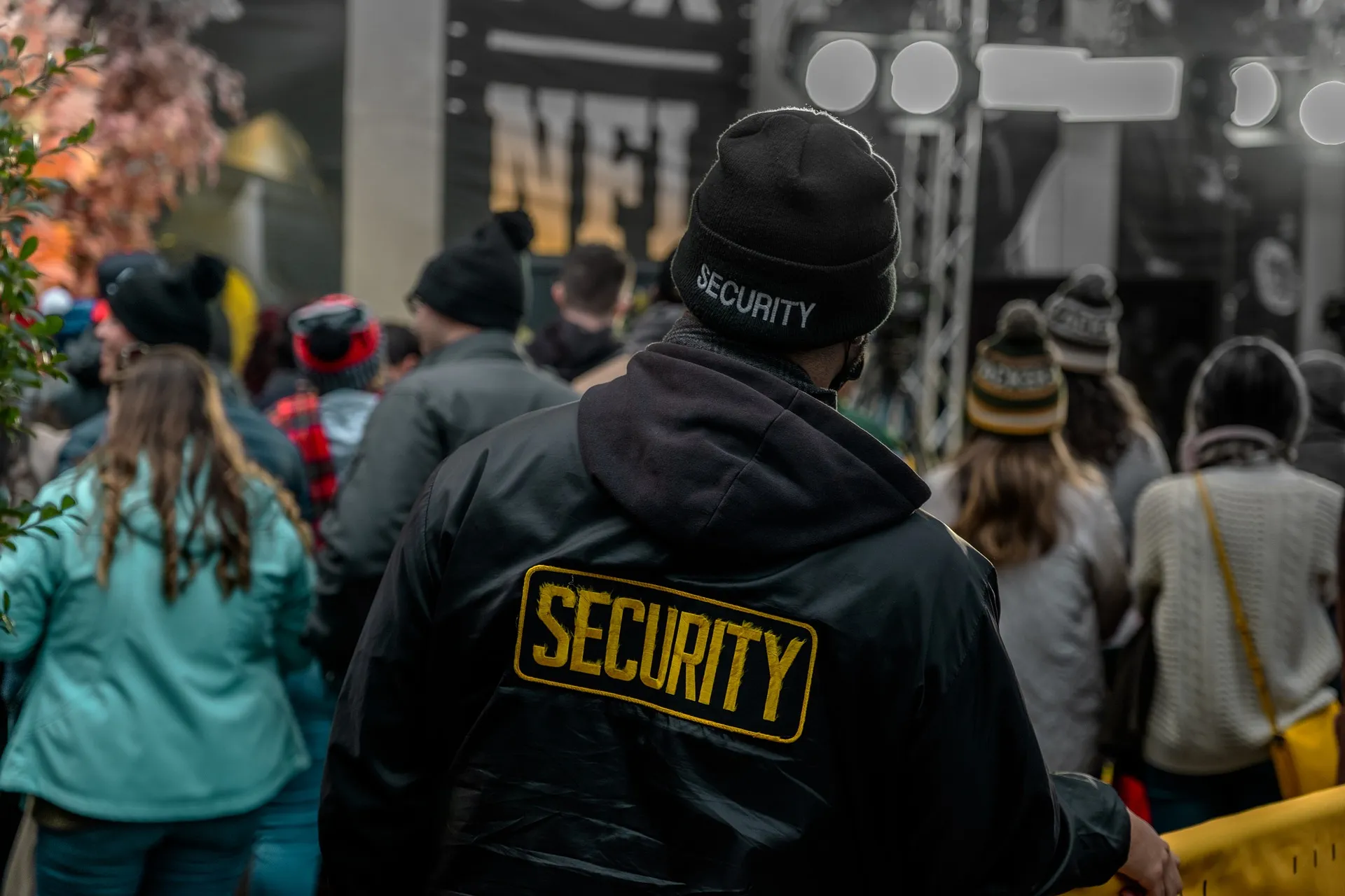 Person in a black SECURITY jacket and beanie seen from behind, facing a crowd in winter hats near a stage with lights