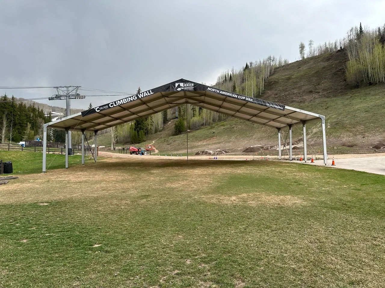 Open-sided frame canopy with banners on a grassy ski-area base below a chairlift and aspen-covered slopes