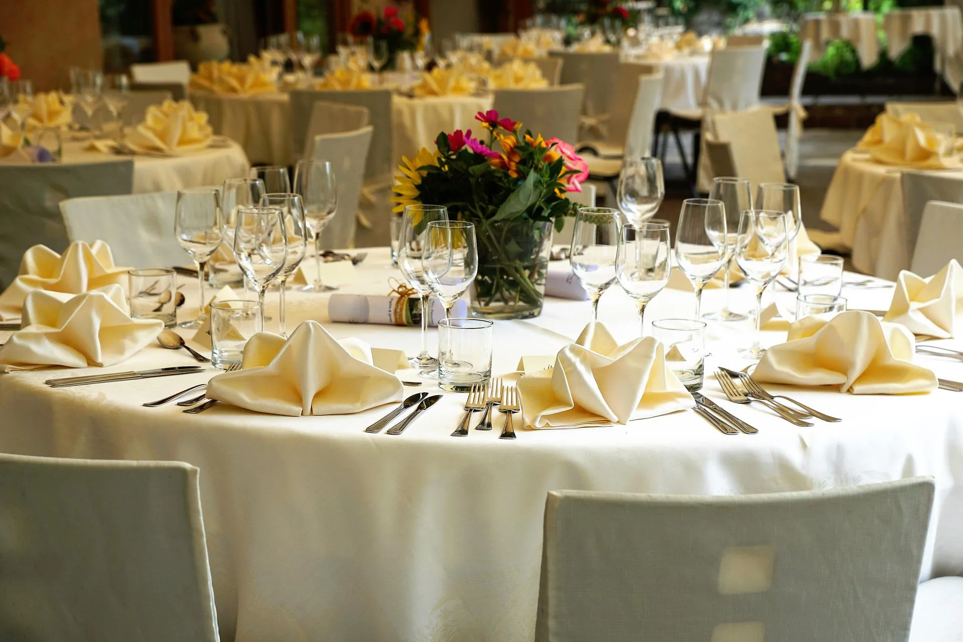 Round table with ivory linen, folded cream napkins, stemware and a bouquet in a glass vase, with covered chairs and more tables behind
