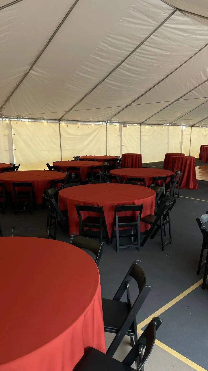 Inside a white tent: round tables with orange-red tablecloths and black folding chairs on gray flooring