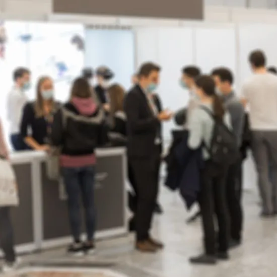 Blurred crowd of attendees, some in face masks, talking around a booth counter in an exhibition hall