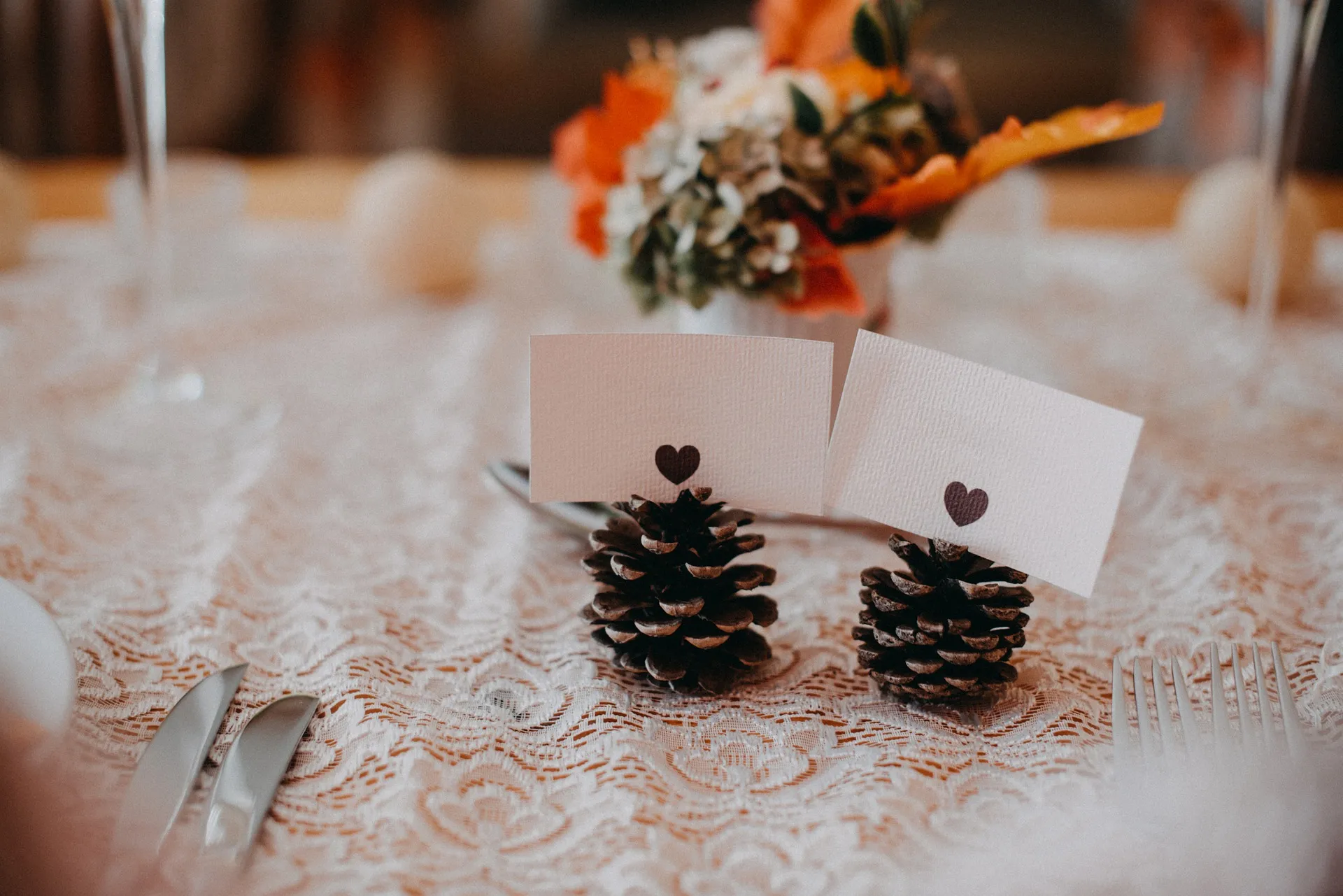 Two heart-marked place cards held in pine cones on a lace tablecloth, with an orange and white flower arrangement behind