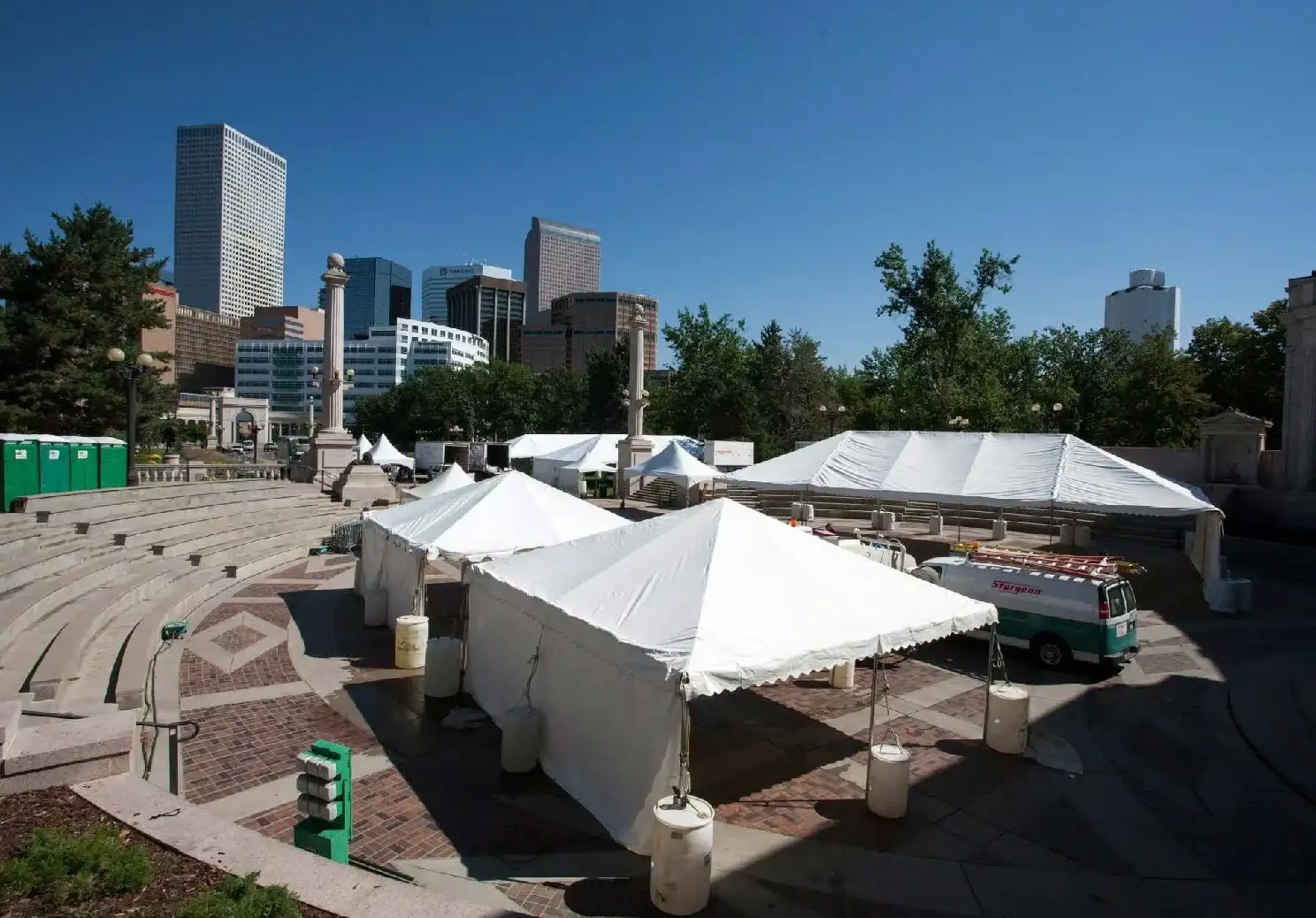 White frame tents weighted with barrels on a brick plaza beside a curved stone amphitheater, with city skyscrapers behind