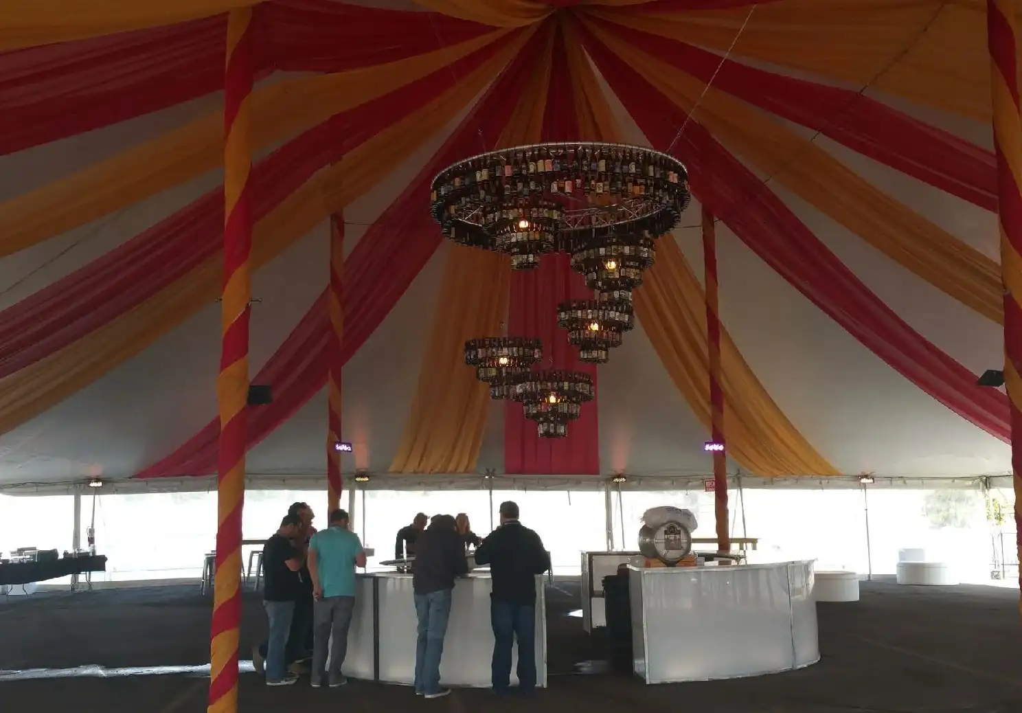 Tent interior with red and gold fabric draped from the center, striped poles, a tiered bottle chandelier and people at white curved bars