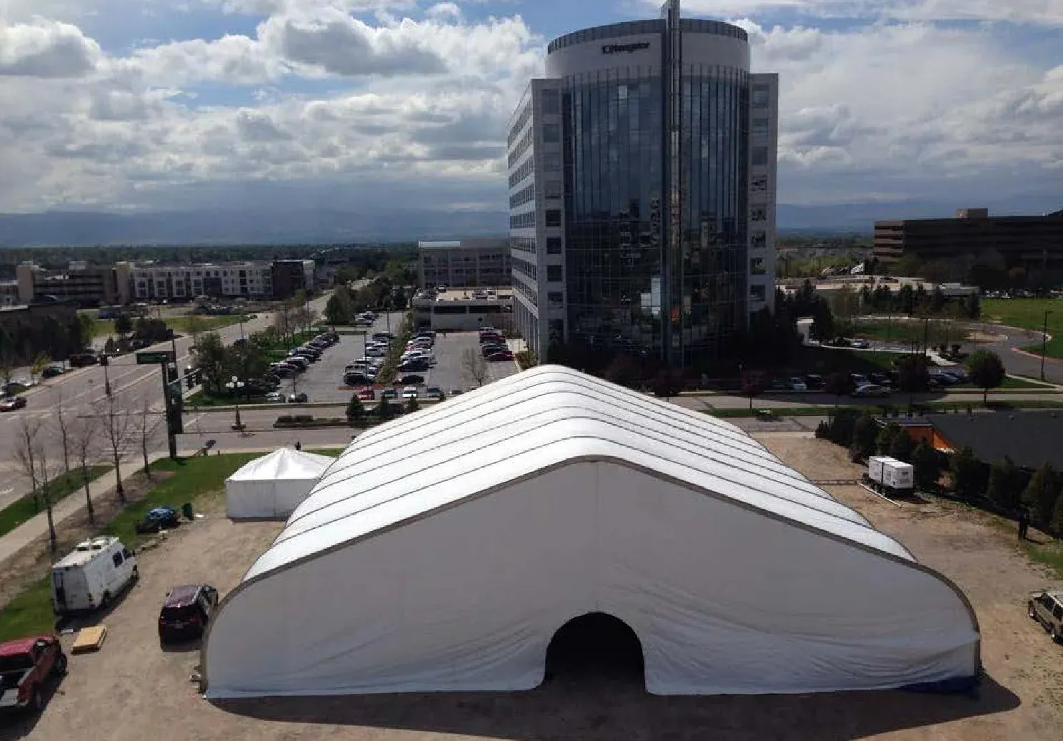 Large white clearspan tent with an arched doorway on a dirt lot, seen from above beside a glass office tower