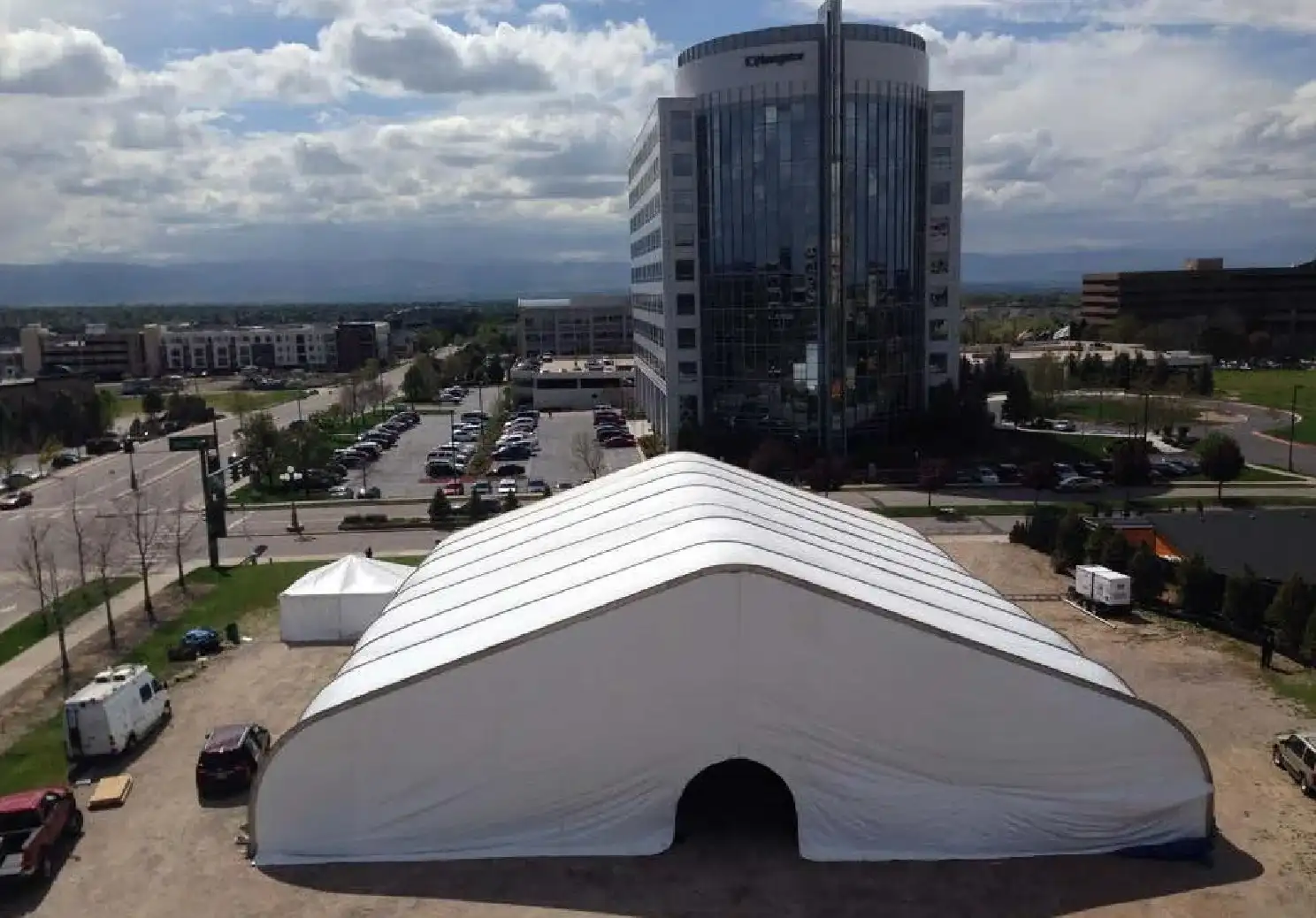 Large white clearspan tent with an arched doorway on a dirt lot, seen from above beside a glass office tower