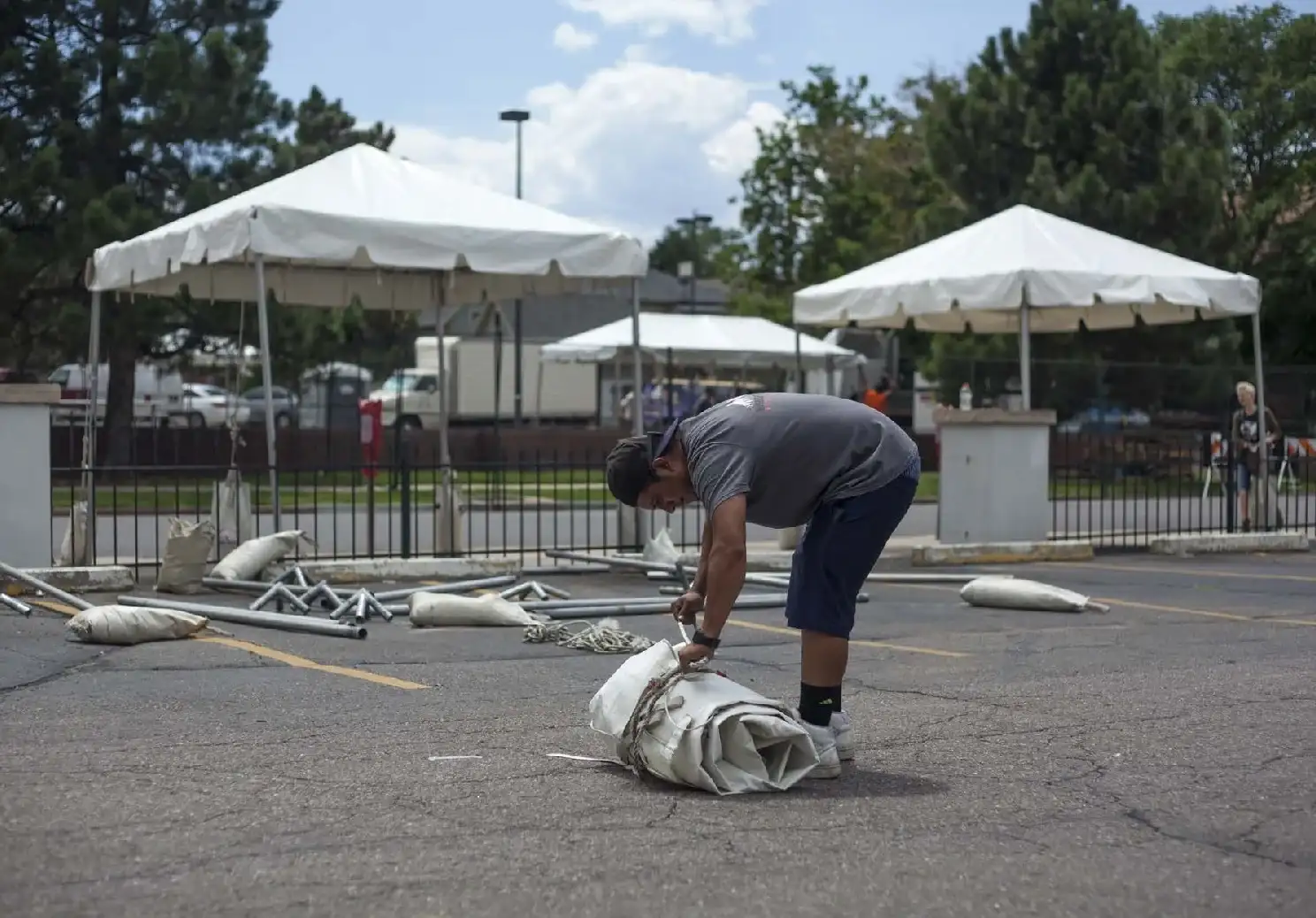 Worker bending over a rolled bundle of tent fabric on a parking lot, with poles and sandbags on the ground and white tents behind