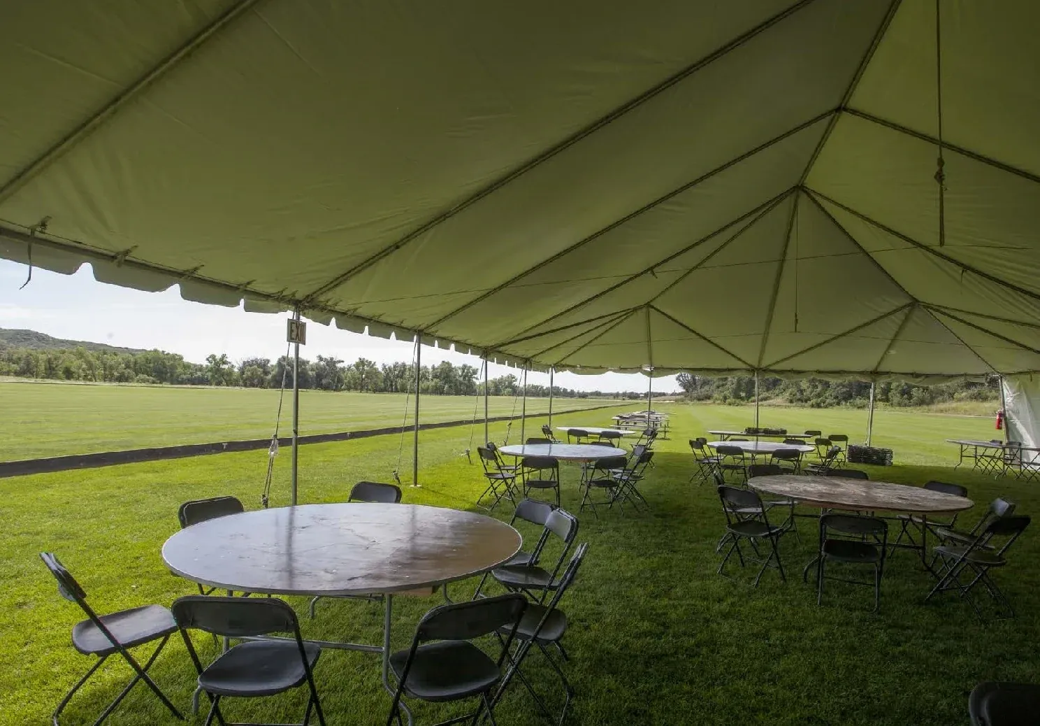 View from inside a white tent over round tables and black folding chairs on the grass, looking out to an open field and trees