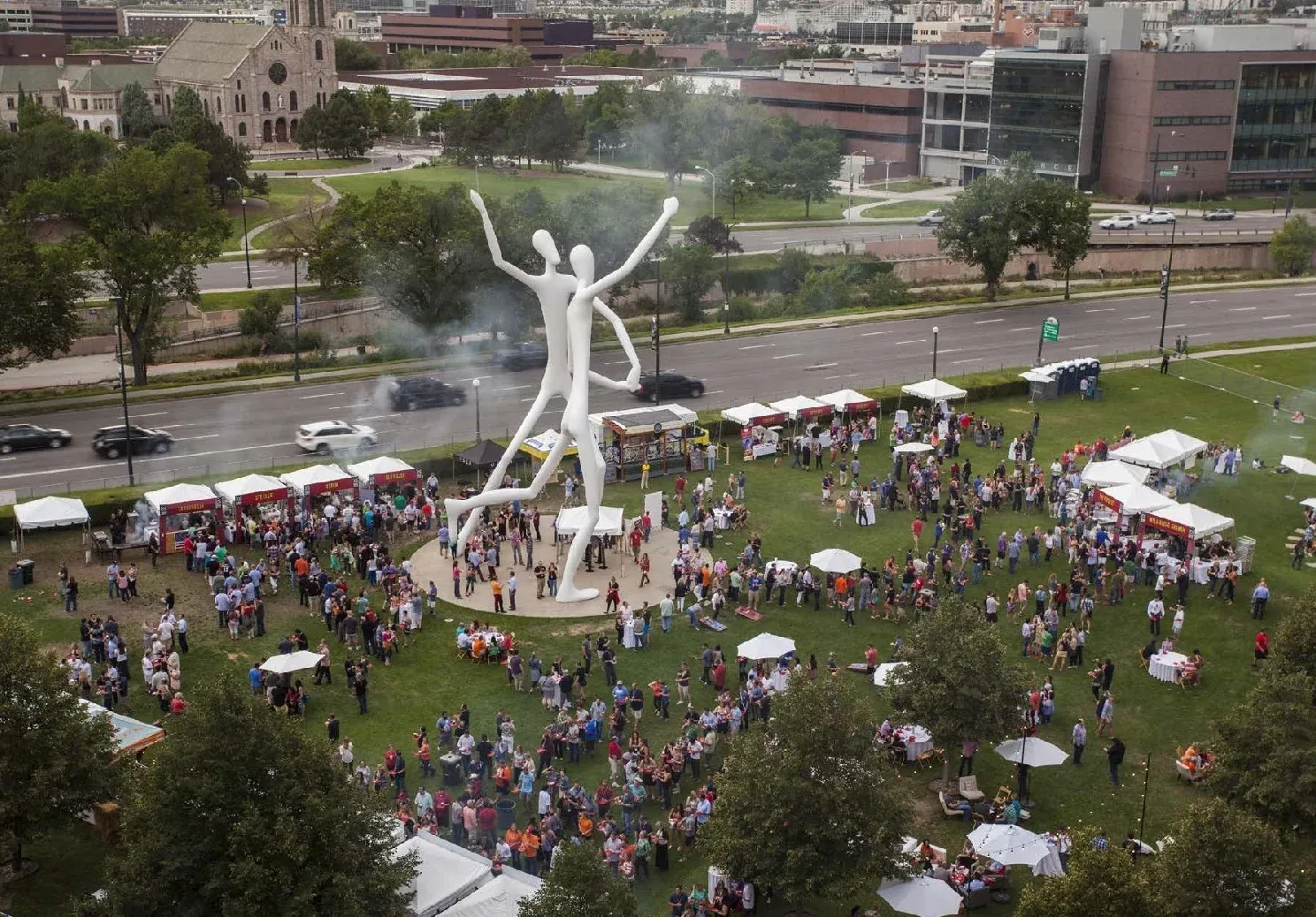 Aerial view of a crowded grass park with small white tents, umbrellas and a giant white two-figure sculpture beside a city road