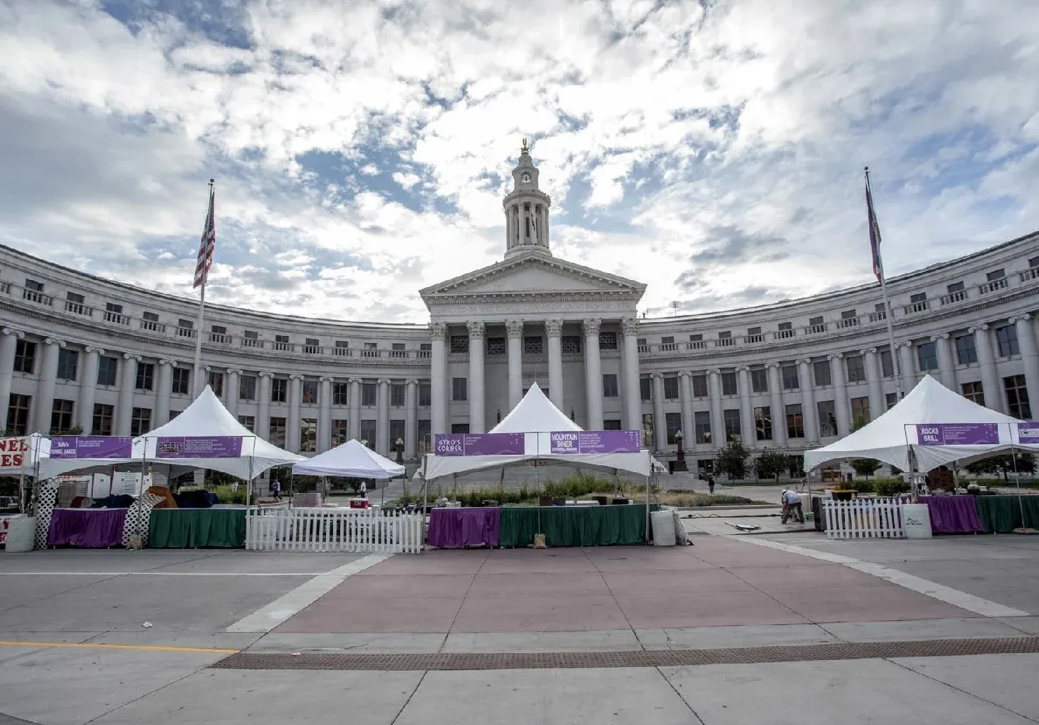 White peaked tents with purple banners, a picket fence and skirted tables on a plaza before a curved columned building under a cloudy sky
