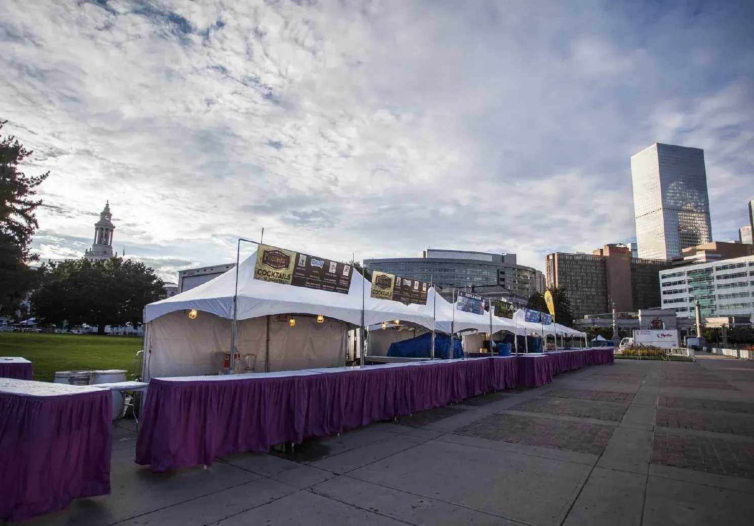 Row of white vendor tents with banners, string lights and long purple-skirted tables along a brick plaza near downtown buildings