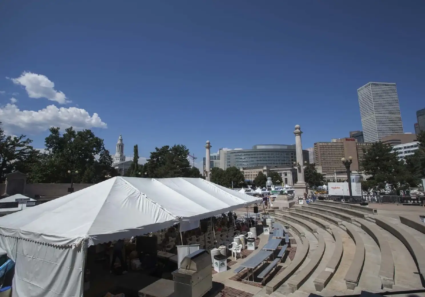 Large white tent with cooking equipment and picnic tables beside curved stone amphitheater steps, with downtown towers under a blue sky