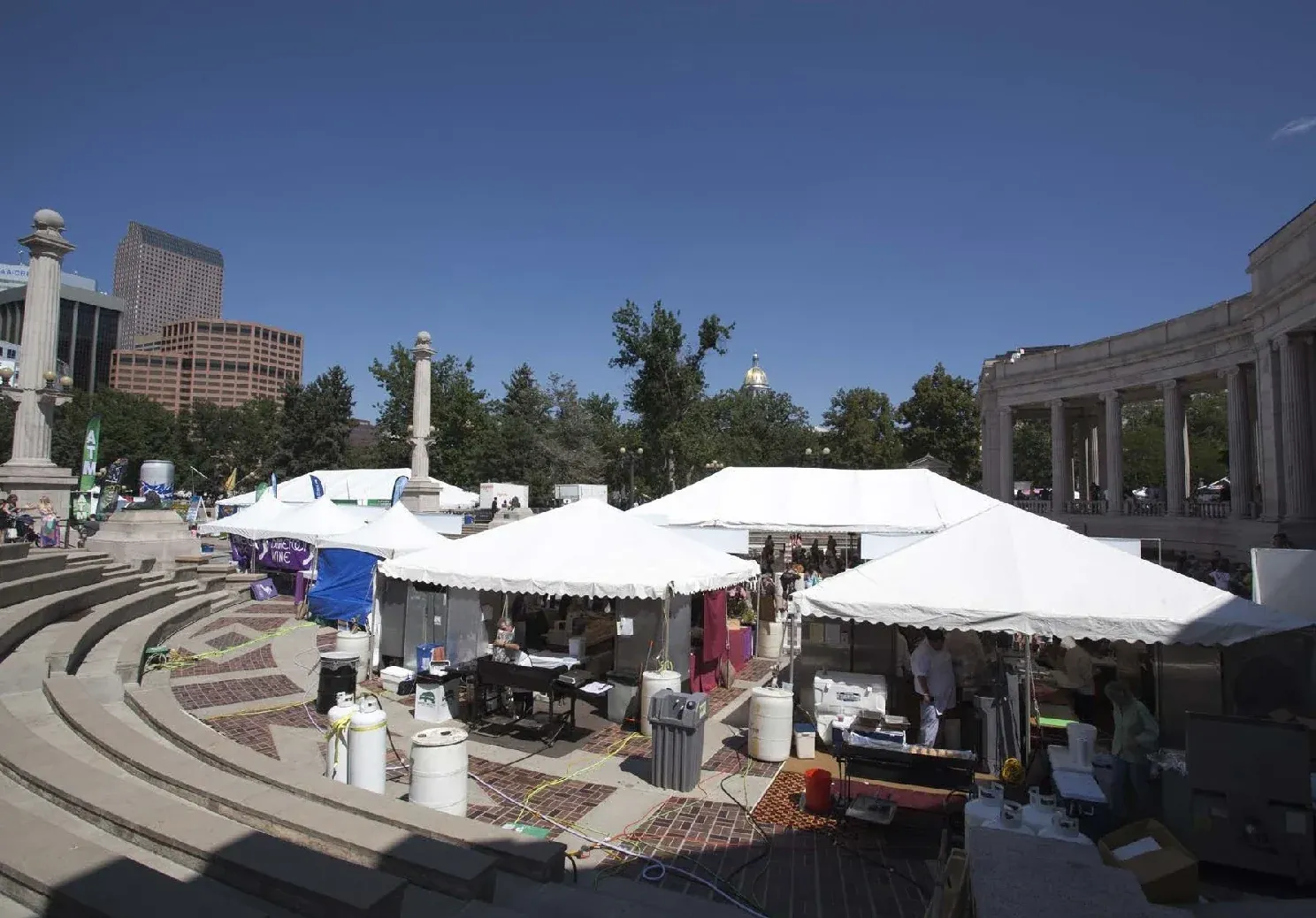 White peaked tents with cooking equipment and propane tanks on a brick plaza below curved stone amphitheater steps, under a blue sky