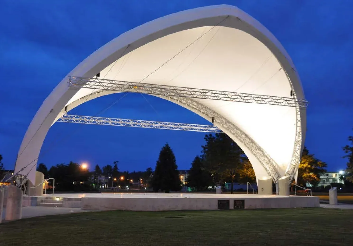 Arched white fabric stage cover with aluminum truss beams, lit at dusk above a concrete stage platform and a lawn