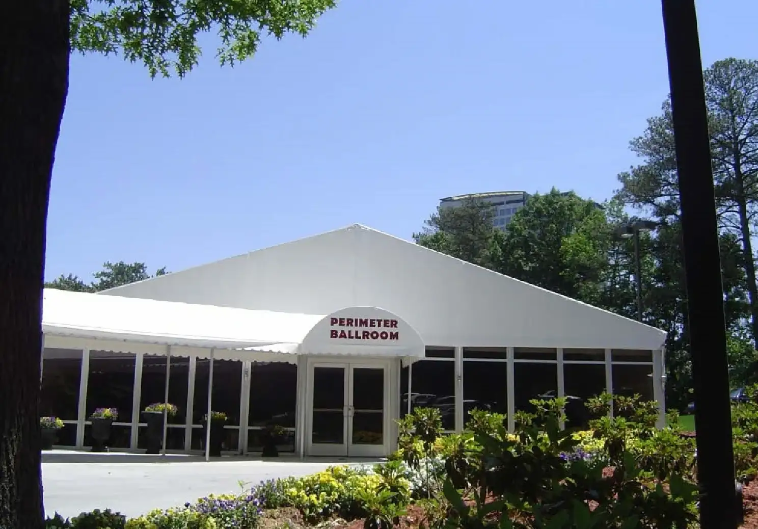 White gable-roof tent with windowed walls, an arched awning over double glass doors, and planters and flowering shrubs in front