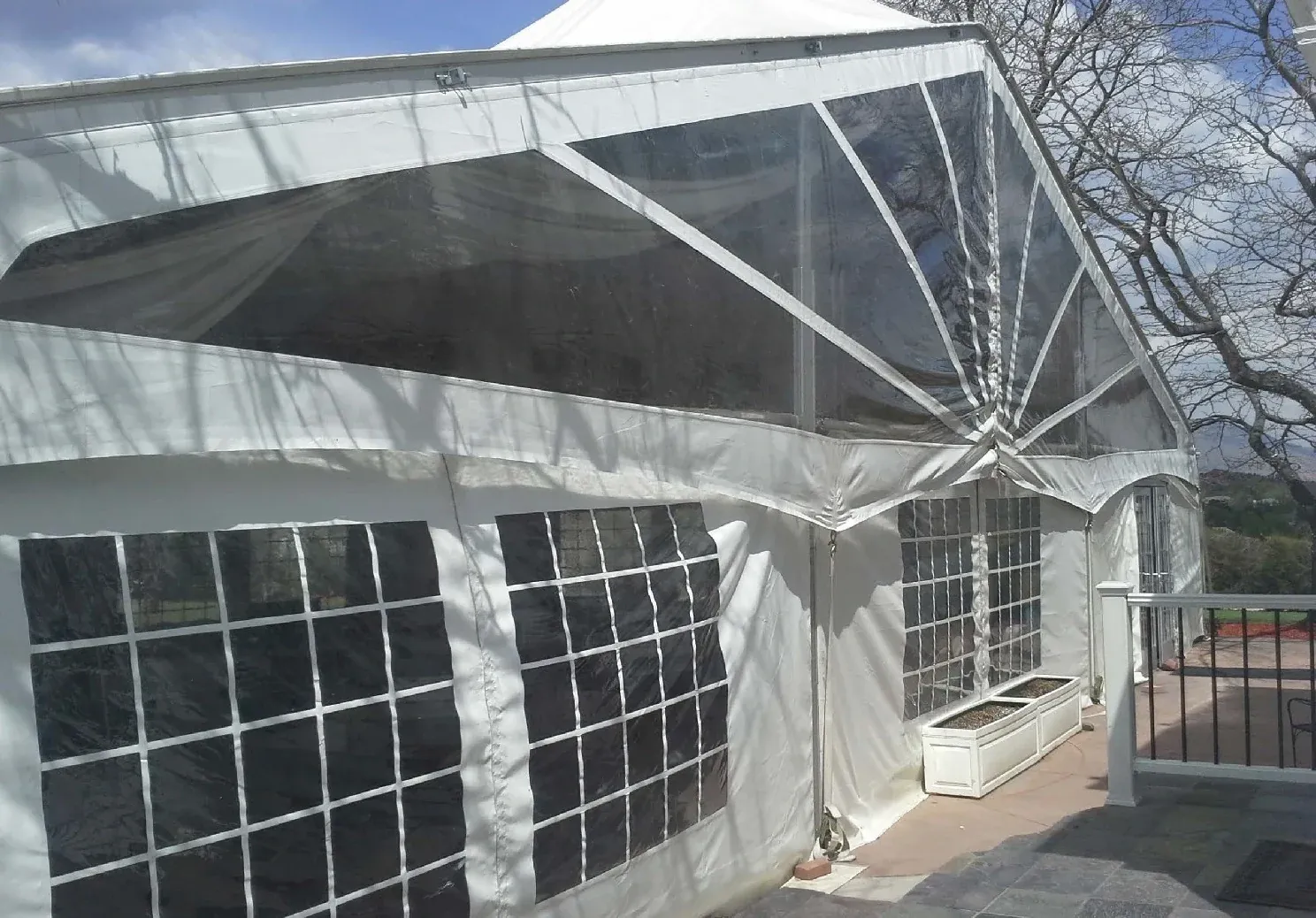 White frame tent with clear upper panels and grid-window sidewalls beside a stone patio, a white deck railing and a planter box