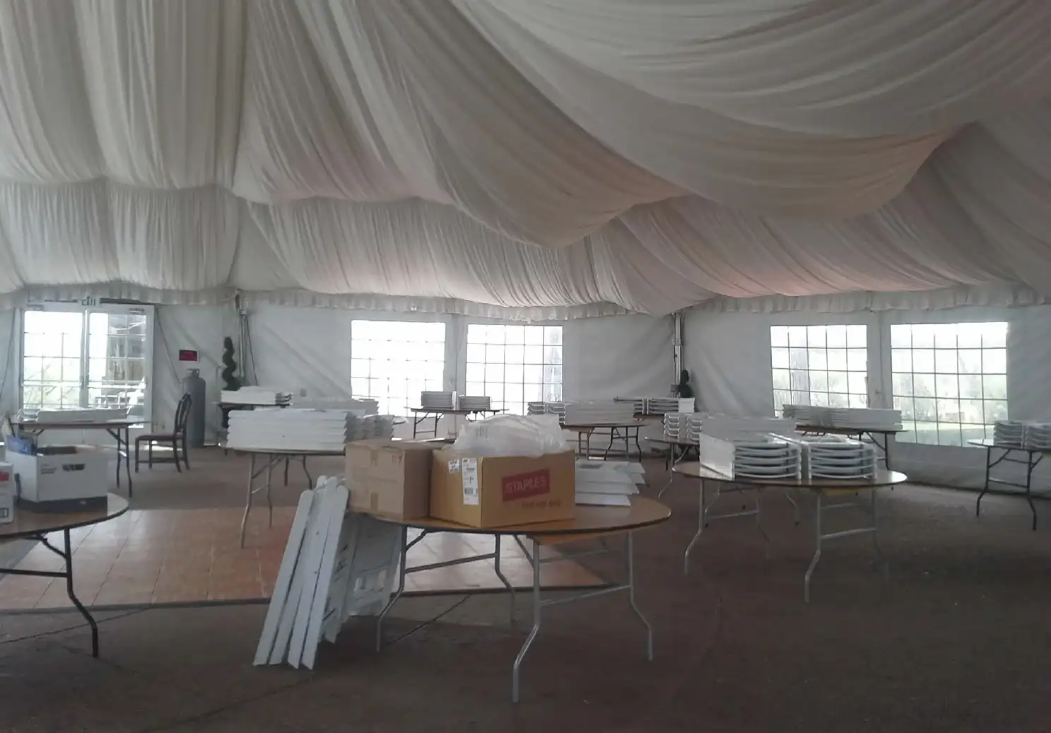 Tent interior with draped white ceiling fabric, window sidewalls, round tables stacked with white chairs and parquet floor panels