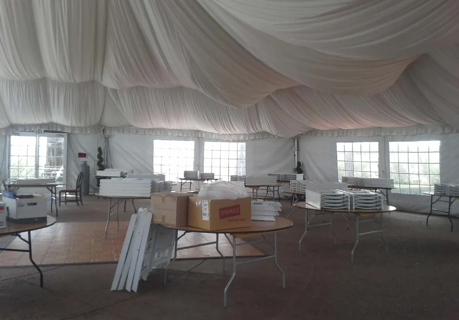Tent interior with draped white ceiling fabric, window sidewalls, round tables stacked with white chairs and parquet floor panels