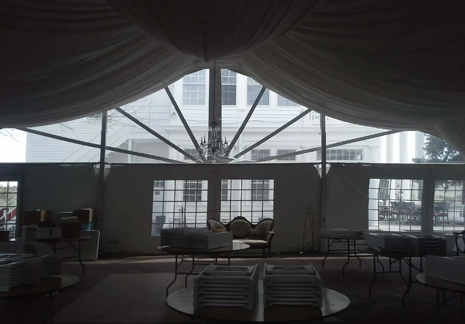 Dim tent interior with draped ceiling fabric, a chandelier, a tufted loveseat and round tables stacked with white chairs