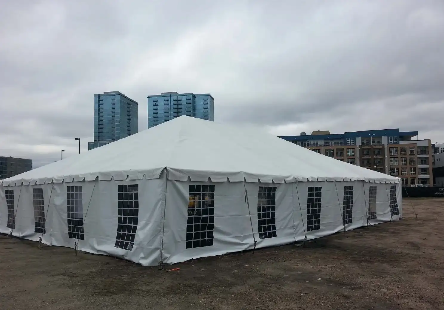 Large white tent with grid-window sidewalls on a bare dirt lot, with glass high-rises and apartment buildings behind under an overcast sky