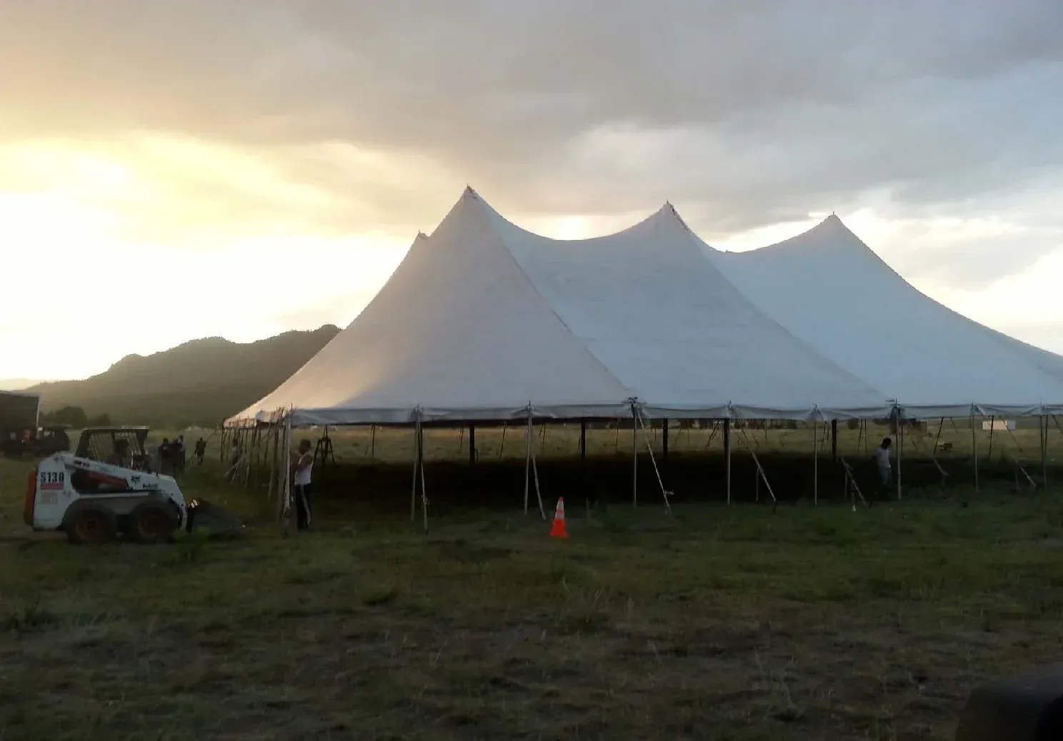 Large white multi-peak pole tent with open sides in a grass field at sunset, with a crew beside it and hills behind