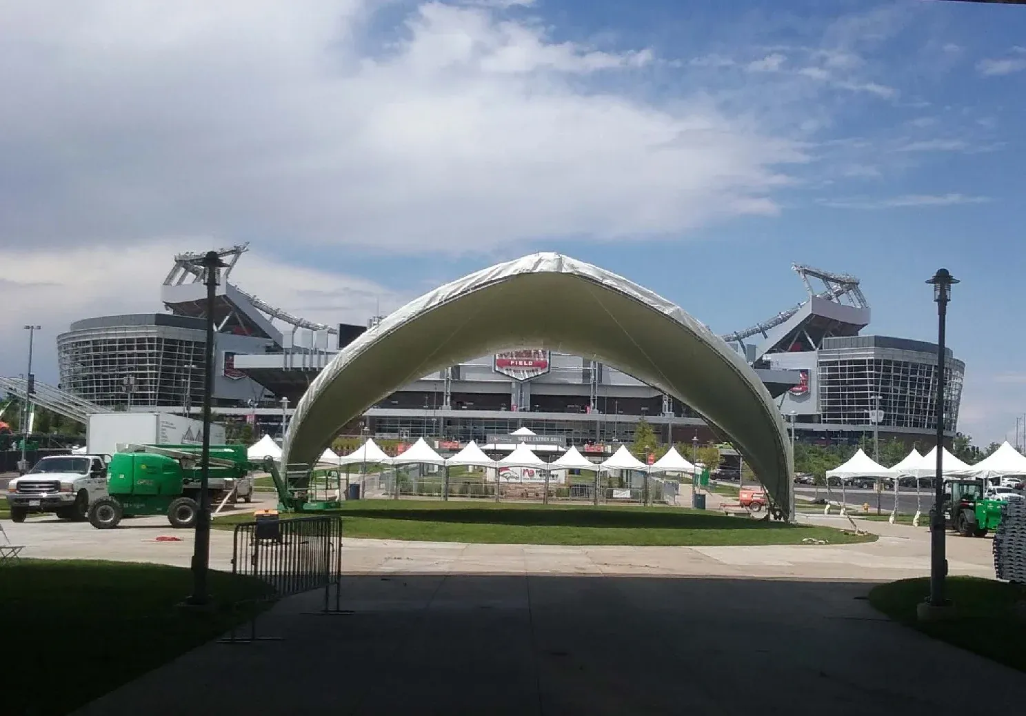 Large white arched fabric canopy on a lawn beside a row of white peaked tents, with a green lift, trucks and a stadium behind